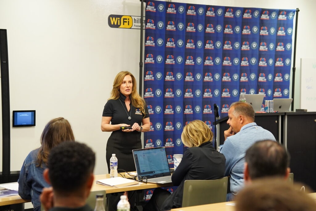 Female corporate executive standing on stage with a clicker presenting operational scaling frameworks to focused business operators at long seminar tables with laptops.