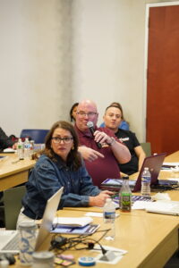 Business operator holding a microphone and pointing while asking a strategic scaling question during an interactive corporate workshop seminar with open laptops on the tables.