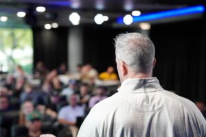 Over-the-shoulder view of a business growth mentor on stage addressing an attentive audience of home service operators during an advanced corporate scaling seminar.