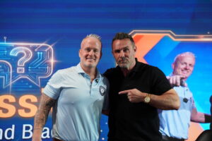 Digital marketing expert Brad Lea in a black polo shirt pointing towards Lance Bachmann on stage in front of a blue presentation screen at a corporate seminar.