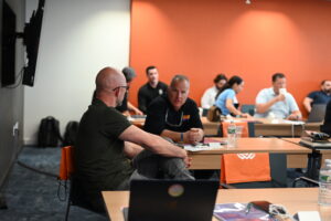 Home service business owners engaged in small group discussion at the Build It to Sell It Mastermind event, seated at tables with orange branded chair bags in the background.