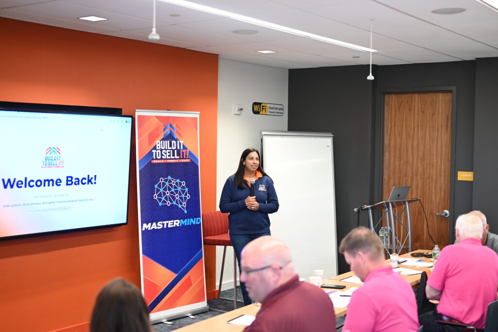 Alicia Johnston speaking at the Build It to Sell It Mastermind event, holding a microphone in front of a branded banner and a "Welcome Back" presentation slide.