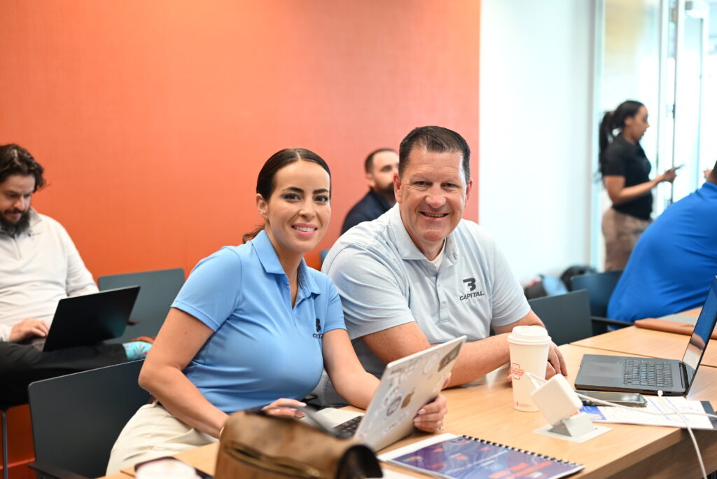 Bill Rossell and Kelly Mesaris of B3 Capital smiling at a mastermind event, seated at a conference table with laptops and other attendees in the background.