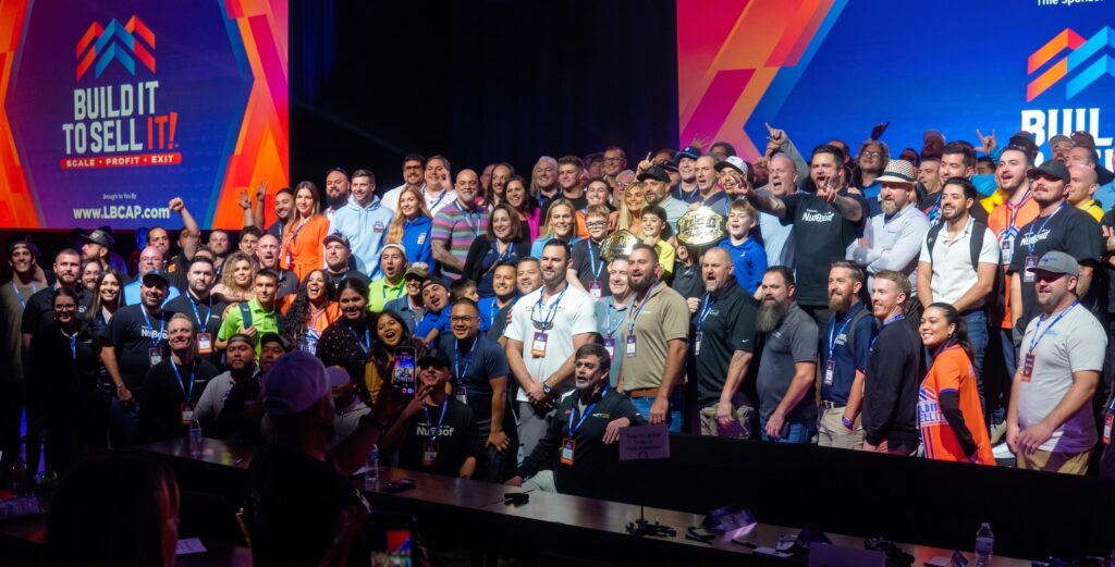 Group photo of attendees at the Build It To Sell It conference standing on stage in front of branded event screens.