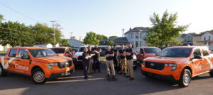 Wide promotional shot of the Tru Choice Roofing team standing in a lot between wrapped orange Ford service trucks for a brand equity feature.