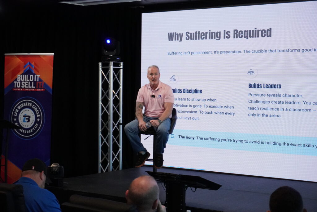 Lance Bachmann speaking on stage during a Winners Circle session with a presentation slide titled “Why Suffering Is Required.”