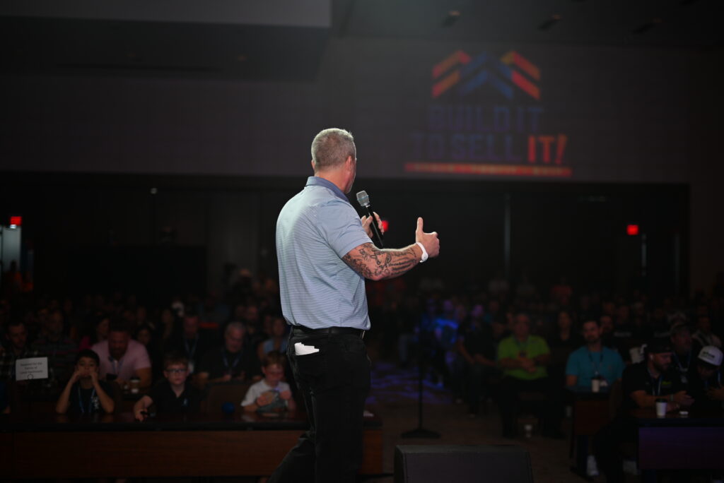 LB Capital founder Lance Bachmann speaking into a microphone on stage in front of a large audience at the official Build It To Sell It Conference.
