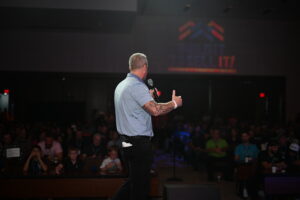 LB Capital founder Lance Bachmann speaking into a microphone on stage in front of a large audience at the official Build It To Sell It Conference.